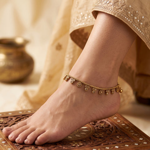 A gold-toned ankle chain bracelet with small dangling charms is shown worn on a person's ankle, with the foot resting on a carved wooden surface and traditional beige fabric in the background.