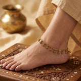 A gold-toned anklet with small dangling coin charms is worn on a person's ankle, shown in a styled close-up with a beige garment and an ornate carved wooden surface.