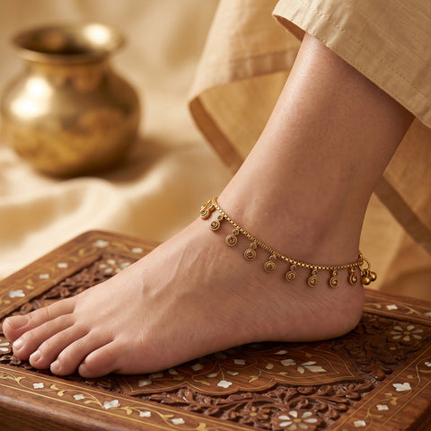 A gold-toned anklet with small dangling coin charms is worn on a person's ankle, shown in a styled close-up with a beige garment and an ornate carved wooden surface.