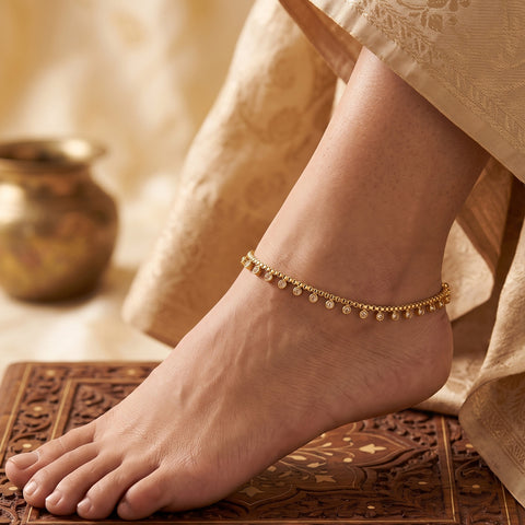 A delicate gold chain anklet with small dangling coins is shown worn on a person's ankle, with the foot resting on an ornate carved wooden surface and draped beige fabric in the background.