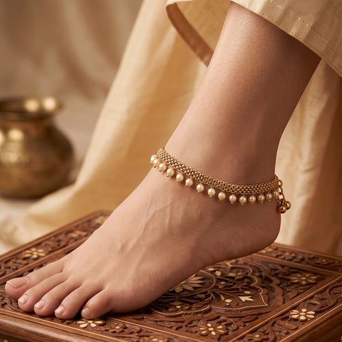 A close-up of a person's foot and ankle wearing a delicate gold-toned chain anklet adorned with small white pearls, posed on a carved wooden surface with draped beige fabric in the background.
