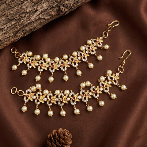 Gold-toned traditional Indian jhumka earrings with white faux pearls, clear rhinestones, and small red floral accents, arranged on a brown satin fabric with a log prop.