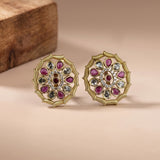 A pair of gold-toned mandala-style stud earrings with pink teardrop-shaped stones, clear round stones, and a central pink stone, displayed on a beige surface with a wooden block in the background.