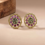 A pair of gold-plated Indian Kundan earrings with purple teardrop-shaped stones, a green stone center, and a gold circular frame, displayed on a beige surface with a wooden block in the background.