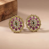A pair of gold-toned Indian Kundan earrings with pink gemstone accents and a central green stone, displayed on a beige surface with a wooden block in the background.