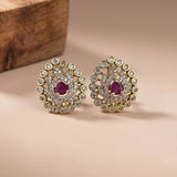 A pair of gold-toned statement earrings featuring oval-cut pinkish-red center stones surrounded by clear pavé-set round stones, displayed on a warm beige surface with a wooden block in the background.