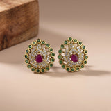 A pair of gold-toned Indian-style stud earrings with central teardrop-shaped ruby gemstones, surrounded by clear pavé stones and a halo of round green stones, displayed on a beige surface with a wooden block in the background.