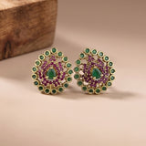 A pair of gold-plated stud earrings featuring a central green teardrop gemstone surrounded by purple stones, with a surrounding halo of green round stones, displayed on a beige surface with a wooden block in the background.