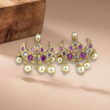 A pair of gold-toned traditional Indian chandbali earrings adorned with purple oval and round stones, clear accent stones, and hanging cream-colored faux pearls, displayed on a soft beige surface with a wooden block in the background.