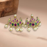 A pair of gold-toned Indian chandbali earrings decorated with green and purple stones and hanging white faux pearls, displayed on a beige surface with a wooden block in the background.