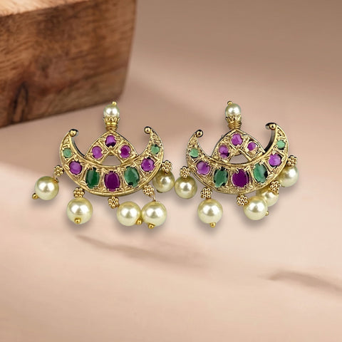 A pair of gold-toned Indian chandbali earrings decorated with green and purple stones and hanging white faux pearls, displayed on a beige surface with a wooden block in the background.