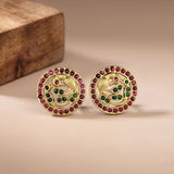 A pair of gold-toned Indian Kundan earrings featuring a peacock motif, embellished with green and pink stones, displayed on a neutral beige surface with a wooden block in the background.