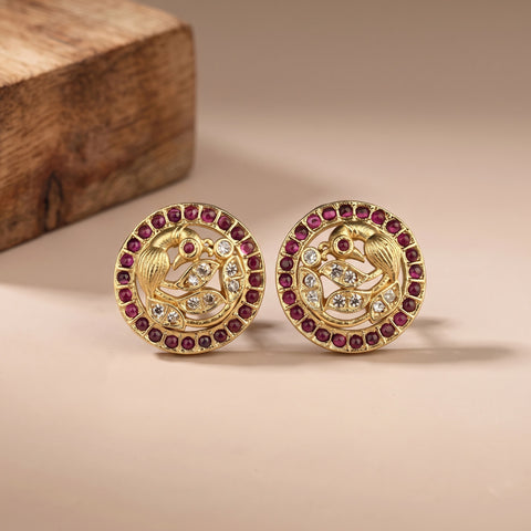 A pair of gold-plated round stud earrings featuring a detailed peacock motif, accented with red gemstones and clear crystals, displayed on a neutral beige surface with a wooden block in the background.