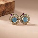 A pair of gold-plated stud earrings featuring round light blue cabochon gemstones surrounded by a halo of small clear stones, with a small purple teardrop gemstone at the top, displayed on a beige surface with a wooden block in the background.