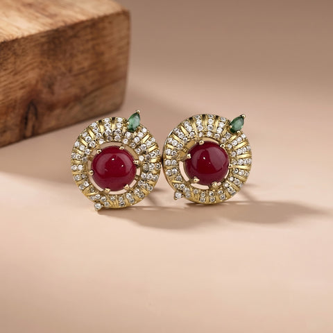 A pair of gold-plated stud earrings featuring a central round deep red cabochon stone surrounded by a halo of small clear crystals, with a small green marquise-cut stone above each. The earrings are displayed on a warm beige surface with a wooden block in the background.