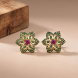 A pair of gold-plated flower-shaped stud earrings decorated with green stones and a central pinkish-red stone, displayed on a beige surface with a wooden block in the background.