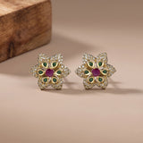 A pair of gold flower-shaped stud earrings with green stone accents, a central pinkish-red stone, and pavé-set clear stones, displayed on a beige surface with a wooden block in the background.
