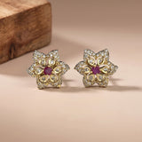 A pair of gold flower-shaped stud earrings with clear pavé stones and a central round purple gemstone, displayed on a warm beige surface with a wooden block in the background.