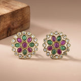 A pair of gold-plated stud earrings with oval green and magenta stones, surrounded by clear round crystals, displayed on a beige surface with a wooden block in the background.