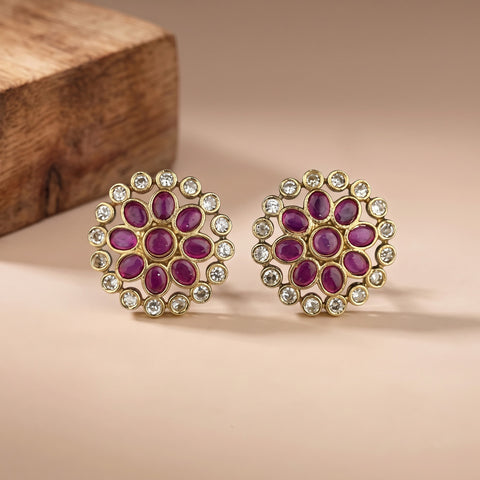 A pair of gold-plated Indian Kundan earrings with oval purple gemstones and clear rhinestones, displayed on a beige surface with a wooden block in the background.