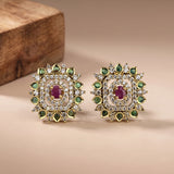 A pair of gold square stud earrings with a central pinkish-red oval stone, surrounded by clear stones and small green stones, displayed on a beige surface with a wooden block in the background.