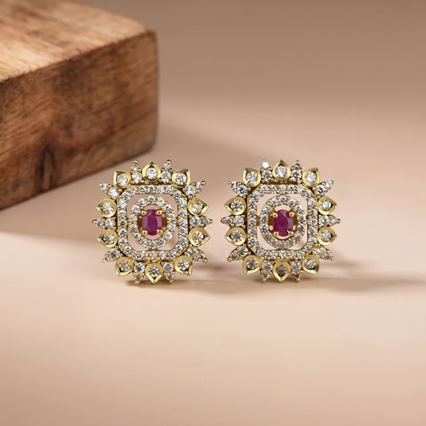 A pair of gold square stud earrings with a central pinkish-red oval stone, surrounded by multiple halo settings of small clear stones, displayed on a beige surface with a wooden block in the background.