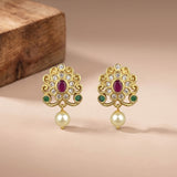 A pair of gold-toned traditional Indian earrings with oval red ruby centers, green stone accents, clear pavé stones, and dangling cream-colored pearls, displayed on a beige surface with a wooden block in the background.