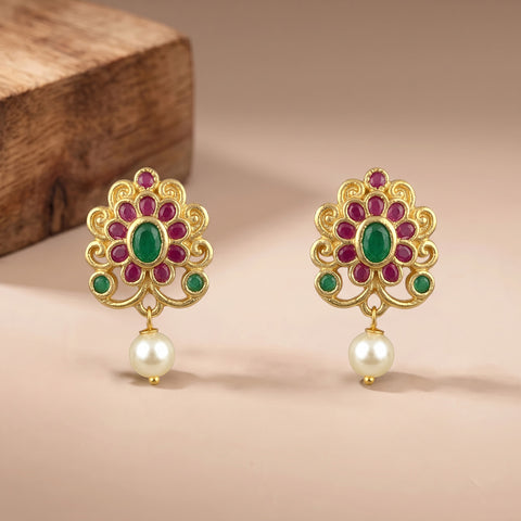 A pair of gold-toned Indian-style earrings with green oval and round stones, pinkish-red petal accents, and dangling white pearls, displayed on a beige surface with a wooden block in the background.