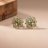 A pair of ornate gold-toned Indian-style floral earrings with green enamel, clear stones, and a small red center stone, displayed on a beige surface with a wooden block in the background.
