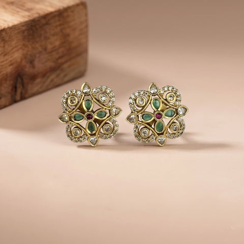 A pair of ornate gold-toned Indian-style floral earrings with green enamel, clear stones, and a small red center stone, displayed on a beige surface with a wooden block in the background.