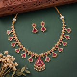 Gold-plated Indian jewelry set featuring a necklace and matching earrings with red inlay and small white bead accents, displayed on a dark green background with decorative dried flowers and a wooden block.