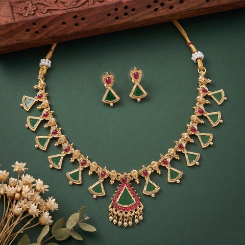 Gold-toned Indian jewelry set featuring a necklace and matching earrings with green and red stone accents, arranged on a dark green background with decorative dried flowers and a carved wooden prop.