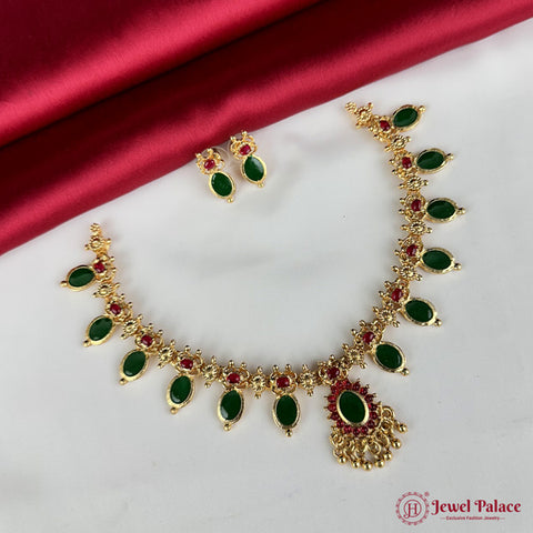 Gold necklace with green and red stones on a white surface with a red fabric background, branded 'Jewel Palace'.