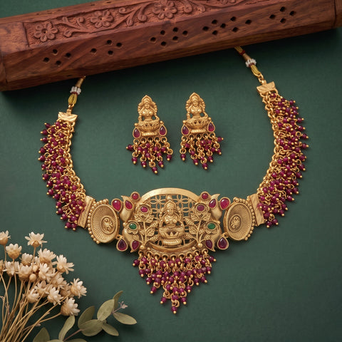 An ornate gold-toned traditional necklace set with matching earrings, decorated with deep red gemstone drops and small green stone accents, arranged on a dark green background with a carved wooden prop and dried flowers.