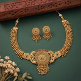 Gold-toned traditional Indian jewelry set featuring a necklace, matching earrings, and a decorative wooden box on a dark green background with small dried flowers in the corner.