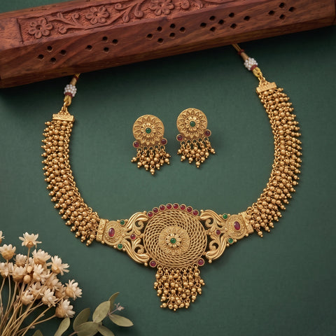 Gold-toned traditional Indian jewelry set featuring a necklace, matching earrings, and a decorative wooden box on a dark green background with small dried flowers in the corner.
