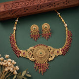 Gold-toned traditional Indian jewelry set featuring a choker necklace, matching earrings, and a small gold pendant, decorated with deep red beaded tassels and green stone accents, displayed on a dark green background with dried flowers and a carved wooden prop.