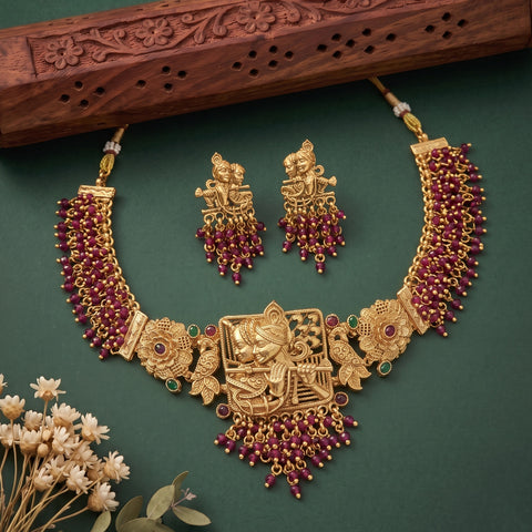 An ornate gold-toned traditional necklace set with matching earrings, decorated with deep red gemstone-like beads and small green stone accents, arranged on a dark green background with a carved wooden prop and dried flowers.