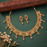 An ornate gold-toned traditional necklace set with matching earrings, decorated with multiple red and green gemstones, displayed on a dark green background with a carved wooden prop and small white flowers.