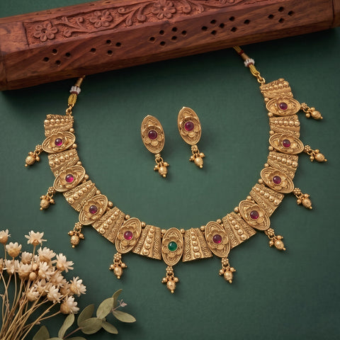 An ornate gold-toned traditional necklace set with matching earrings, decorated with multiple red and green gemstones, displayed on a dark green background with a carved wooden prop and small white flowers.