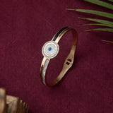 A rose gold bangle bracelet with a circular white mother-of-pearl face, a blue evil eye, and a border of clear crystals, displayed on a textured burgundy fabric background with green leaves.