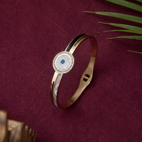 A rose gold bangle bracelet with a circular white mother-of-pearl face, a blue evil eye, and a border of clear crystals, displayed on a textured burgundy fabric background with green leaves.