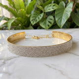 A wide gold-toned choker bracelet covered in rows of clear rhinestones, resting on a white marble surface with green plants blurred in the background.