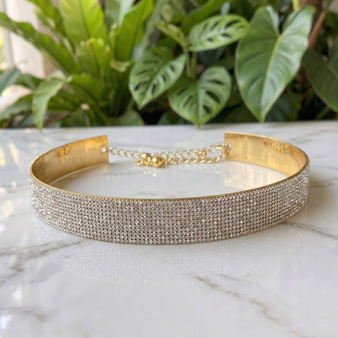 A wide gold-toned choker bracelet covered in rows of clear rhinestones, resting on a white marble surface with green plants blurred in the background.