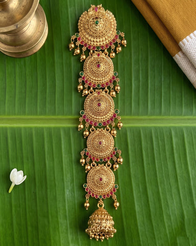 An ornate gold-toned traditional Indian jewelry piece with five stacked circular motifs, red and green stone accents, dangling gold bells, and a large bell-shaped pendant at the bottom, displayed on a green banana leaf with a brass vessel and fabric in the background.