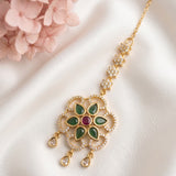 Gold-toned Indian bridal jewelry piece featuring a detailed floral pendant with green enamel leaves, a central red stone, and clear stones, displayed on a white satin fabric with pink flowers in the corner.