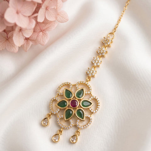 Gold-toned Indian bridal jewelry piece featuring a detailed floral pendant with green enamel leaves, a central red stone, and clear stones, displayed on a white satin fabric with pink flowers in the corner.