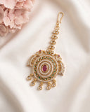 An ornate gold-toned traditional Indian jhumka earring decorated with white stones, green stones, and a central pink stone, displayed on a draped white fabric with pink flowers in the corner.