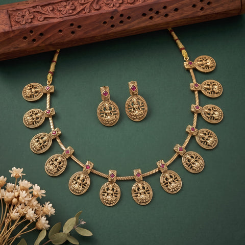 Gold-toned traditional necklace and matching earrings with circular deity motifs and small red stone accents, arranged on a dark green background with dried flowers and a carved wooden prop.