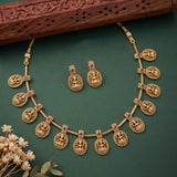 Gold-toned traditional jewelry set featuring a necklace and matching earrings with circular carved motifs and small red and green stone accents, arranged on a dark green background with a carved wooden prop and small dried flowers.